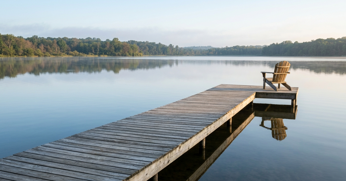 Wooden dock extending into calm lake with Adirondack chair, forested shoreline reflected in still water