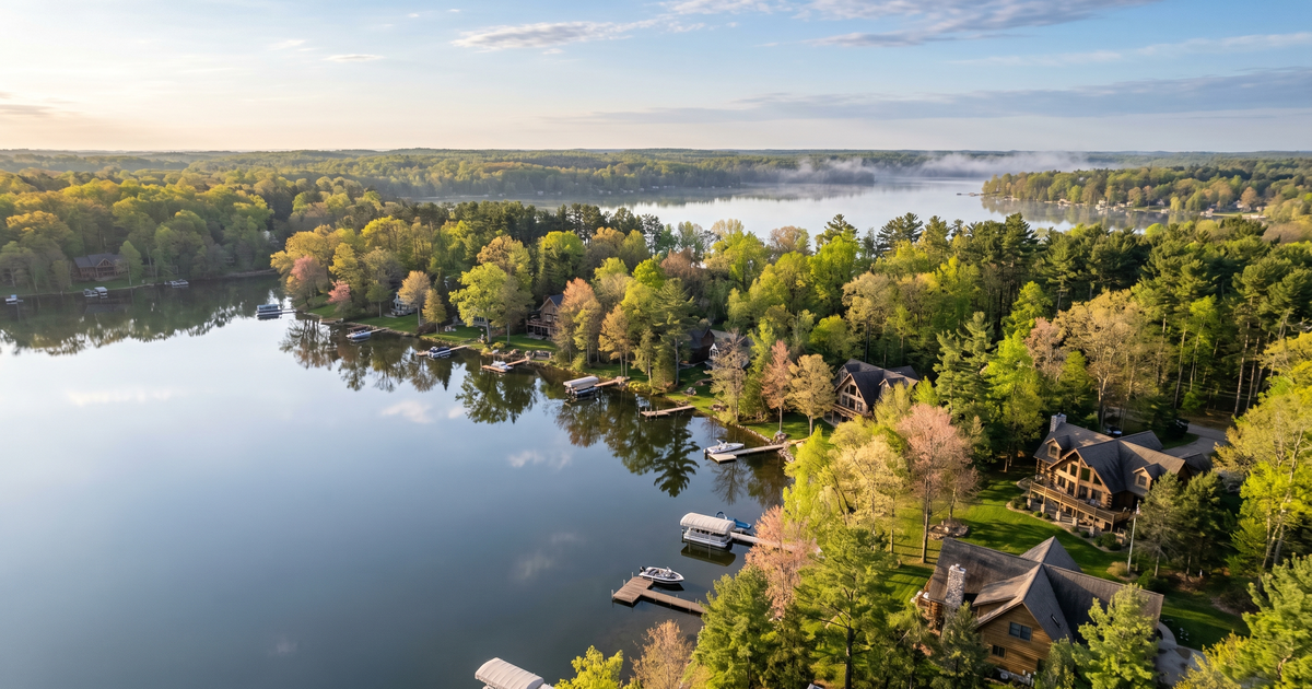 Aerial view of a Southwest Michigan inland lake on a calm spring morning with lakefront homes, private docks, and mirror flat water reflecting the shoreline trees.